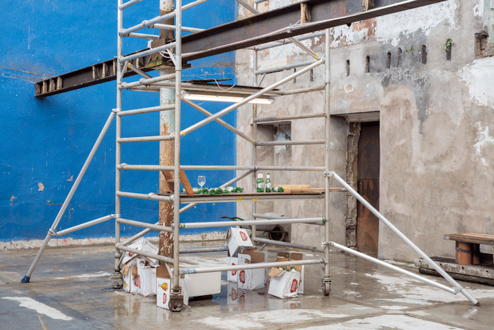 A metal scaffolding with beers of Stella Artois and wine glasses scattered on it. Below it the cardboxes of beer. A blue wall behind it, a cement wall to the right.