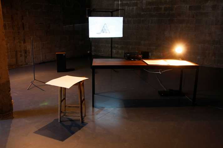 rustic room with brick walls, the lighting is low. in the foreground there is a wooden table and stool. in the background there is a small screen that an image of a tower of cards is being projected onto.