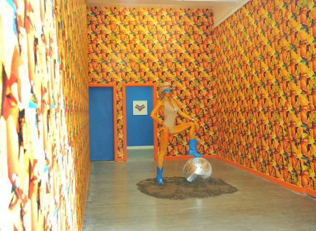 mannequin in a swimsuit and a straw hat standing with one foot on a disco ball in a pile of dirt. the room is covered in bright patterned yellow wallpaper