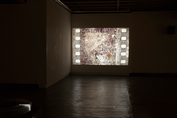 A dark, large room with rough flooring and plain walls, the ceiling has iron beams running across it. The wall in the front has an image of an old film reel style formatted image of something abstract.