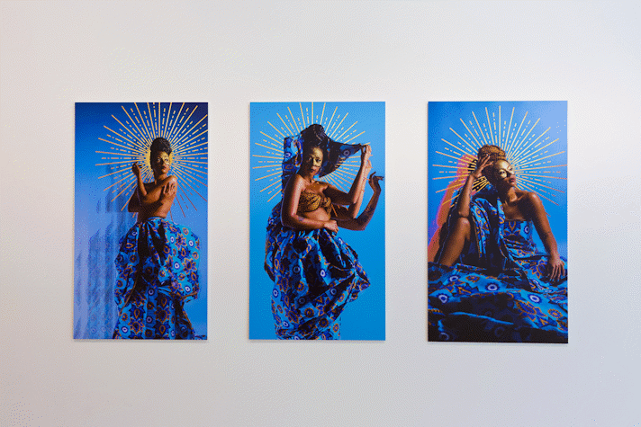 A series of portrait photographs of people of colour on bright blue backgrounds with abstract shapes around their heads, hang on a gallery wall