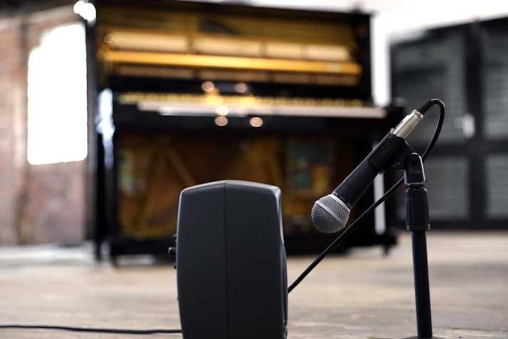 The image shows Yuko Mohri's installation in the pipe factory. In the foreground is a small black speaker. A microphone is pointed toward the speaker. In the background, blurred, is an upright piano.