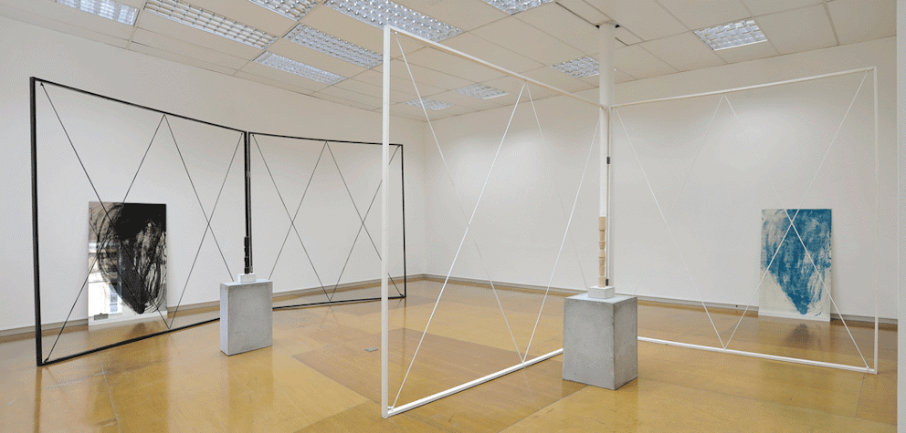 gallery installation. In the back left of the image there is a mirror with black abstract markings leaning against the wall. in front is a black metal frame with criss-crossing wire forming 4 X's. In front of this is a small concrete plinth with a stacked black sculpture atop. the far right shows a white canvas leaning against the wall with abstract blue markings. in front of this is a white frame identical to the black one, with 4 wire X's crossing it with another concrete plinth with small sculpture atop.