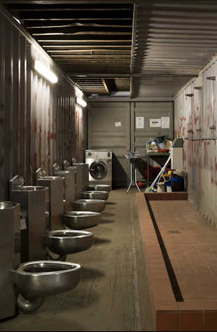 a narrow room with stainless steel urinals, a tiled shower area, and a washing machine surrounded by cleaning supplies. The walls and ceiling are constructed from metal shipping container panels.