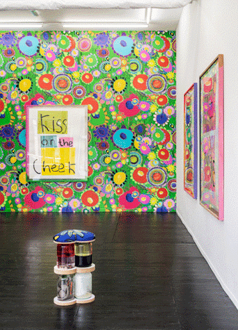 The image shows a colourful exhibition with bright patterned wallpaper and sculptures made of glass jars