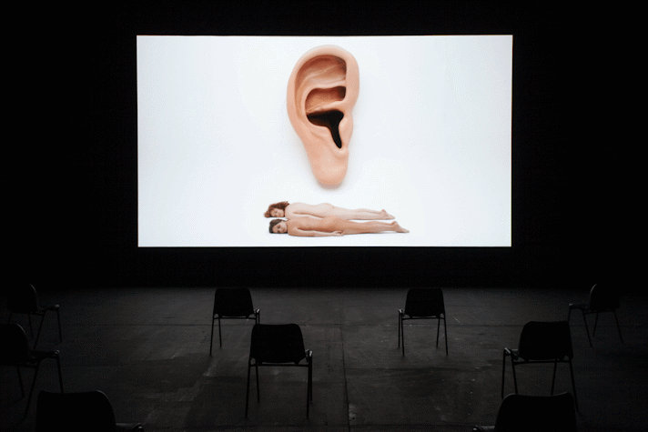 image of film being screened in large dark room. the screen shows a giant ear and two naked bodies lying one atop the other.