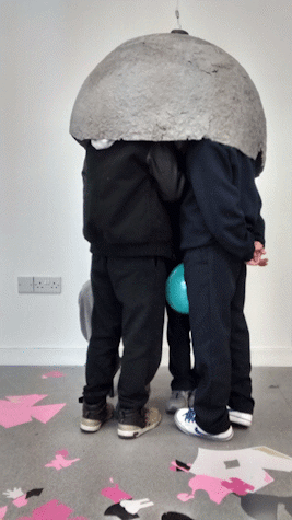 A number of figures stand close together in a gallery space. Their heads are all hidden inside a grey dome shape suspended form the ceiling. Pink and black cut out paper shapes are lying n the floor.