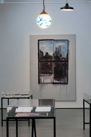 large white canvas with a painting in the centre of the view from a window. in front there are black metal tables with glass encasing. there are notebooks beneath the glass.