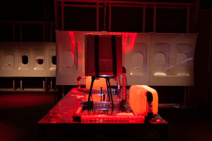Image of Jenkin van Zyl's installation at Tramway. The room sits in eerie red light with strange panels of an aeroplane, a watertank, pipework and fridge.