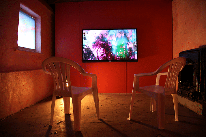 A room with orange walls and a small light, with two white plastic chairs looking at a screen. The screen depicts some trees with pink, blue and green colours. There is a black heater to the right, and a blue printed vynil to in the left wall.