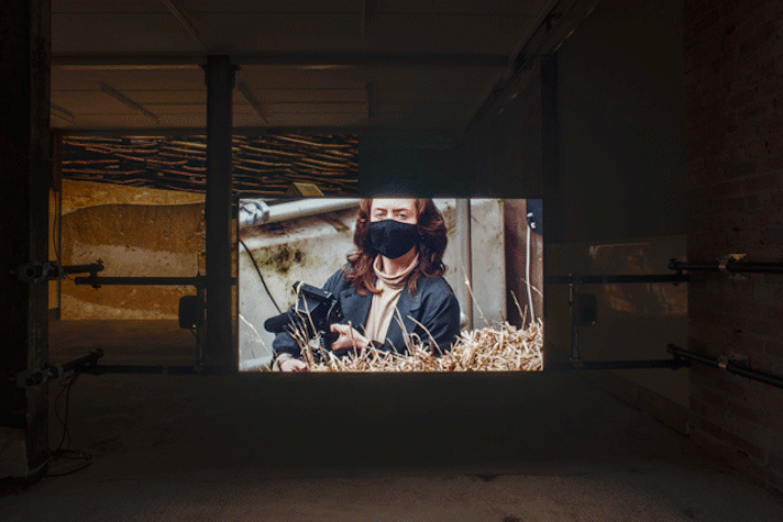 a dark industrial space with film projected. On the screen is a light-skinned person with long brown hair, a black facemask, holding a camera. In the foreground is a bale of hay