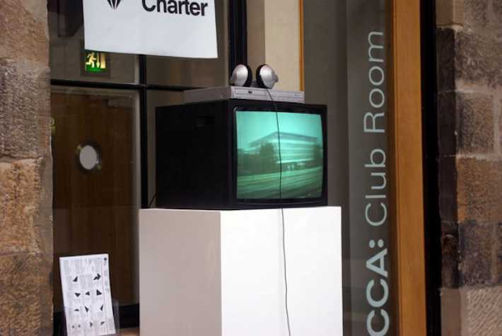 cube monitor atop a plinth. in the background text on the wall reads 'CCA: Club Room'.