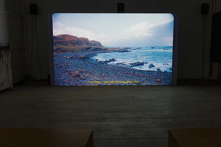 There is a bright screen in a dark room, with film projected onto it. The scene is of a rocky beach.