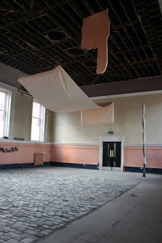 Installation view of a large room with a cobbled floor, a double door at the back and section of material suspended from the ceiling. There are windows also visible.