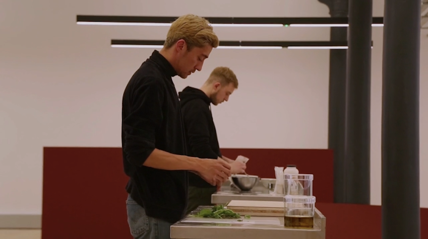 Two white men under fluorescent lights are wearing a black t-shirt and are mixing things like a in a lab experiment. There is a red panel behind them.