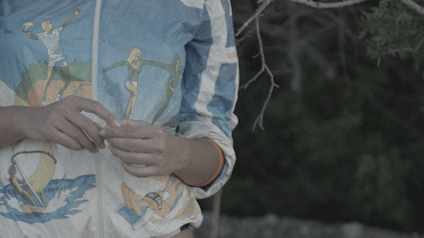 Film still showing the middle part of a figure (torso) wearing a jacket with hands held in front. Trees behind.