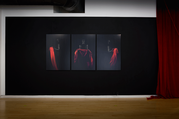 on a black wall at a distance three portrait photographs hang, showing a black figure on a black background holding a bright red drape of fabric