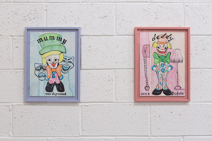 Two colour pencil drawings hang on a white blockwork wall. On the left, in a pale blue frame the drawing shows a clown and black text that reads 'mummy was depressed'. On the right in a salmon pink frame there is a clown and black cursive text that reads 'daddy was a trickster'