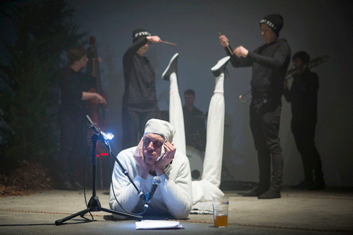 In a dark room, the spotlight is on a man, dressed in all white, lying on the floor with his head propped up on this hands. He has extremely long legs. There is a microphone in front of his face, a sheet of paper below it and a glass of some drink on his right. Behind him are five people, all dressed in black, playing instruments. On the right is a man playing the cello, drums at the back and the saxophone on the left side.