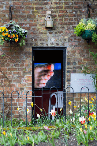 A brick building with flowers in the garden and on the sides of the door. Through the door we can see a video of painted toenails and a white plastic chair. To the right of the open door we can see the title and authors of the exbition in a white sign, pink lettering.