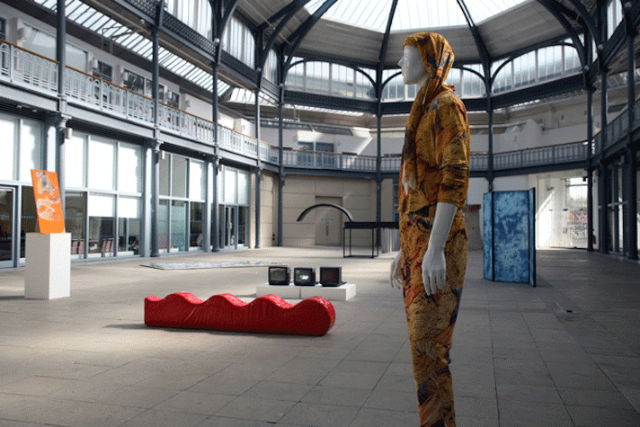 mannequin wearing a suit with images of pasta on it. behind there is a red curvy bench like structure on the floor, three cube monitors on a low white stand and various other scultpural artworks.