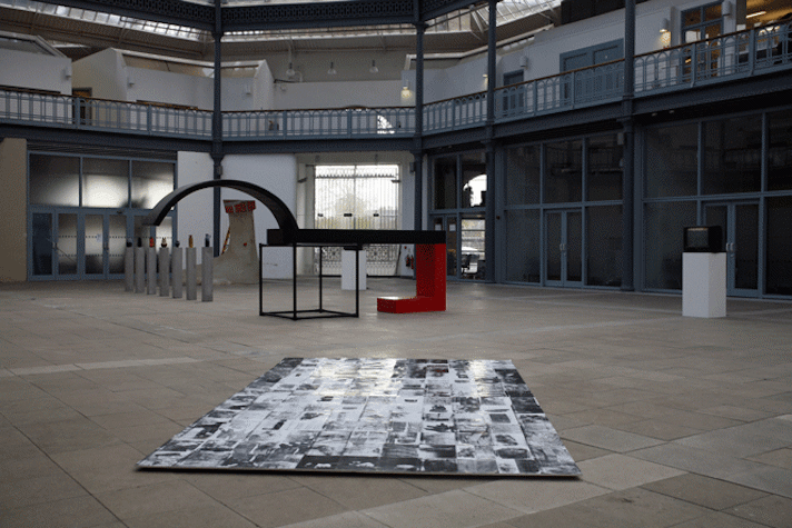 multiple artworks in a large market hall. in the foreground, on the floor, there is a large flat collage of black and white photos. in the background there are various geometric sculptures and a row of concrete plinths with ceramic vases atop.