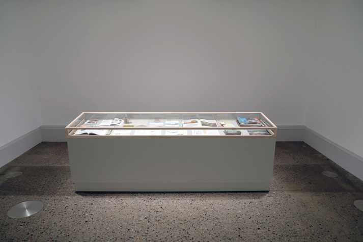 A glass vitrine sits on a large white plinth. Inside the vitrine are a collection of open sketchbooks and books.