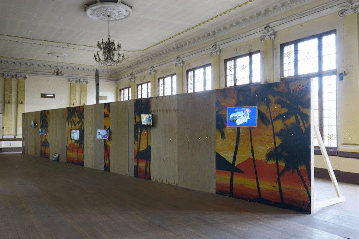 A plywood wall with segments of a sunset in the beach and plain plywood with screens with video in Kelvin Hall as a part of Glasgow International 2018.