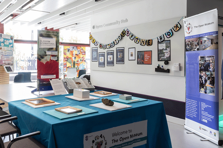 A well-lit room has art displayed on the walls and items displayed on a table which is covered by a blue tablecloth.