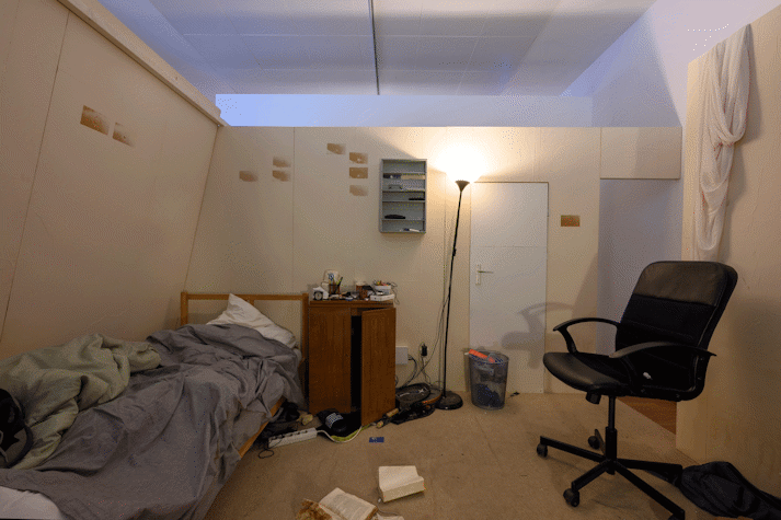 A bedroom space with white walls filled with a bed, a computer chair and storage furniture. On the floors sits open books and cables.