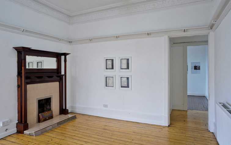 room with wooden floors in a traditional tenement building, left wall has a wood and sandstone fireplace. back wall has 4 framed prints arranged in a square
