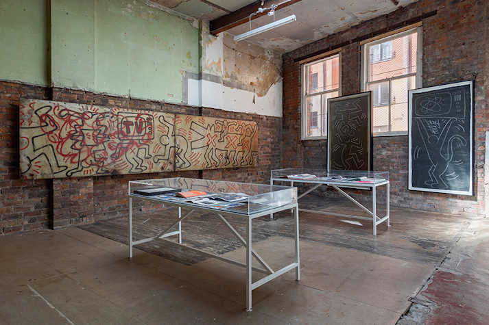 Artwork is displayed on brick walls in an industrial space. In the centre are are two glass-topped tables.