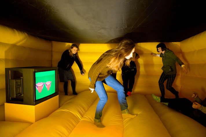 In a bouncy castle, four people are jumping around. There is a small monitor in the middle of the left side wall of the bouncy castle. The monitor has an image of two mouths sticking their tongues out in a bright background.
