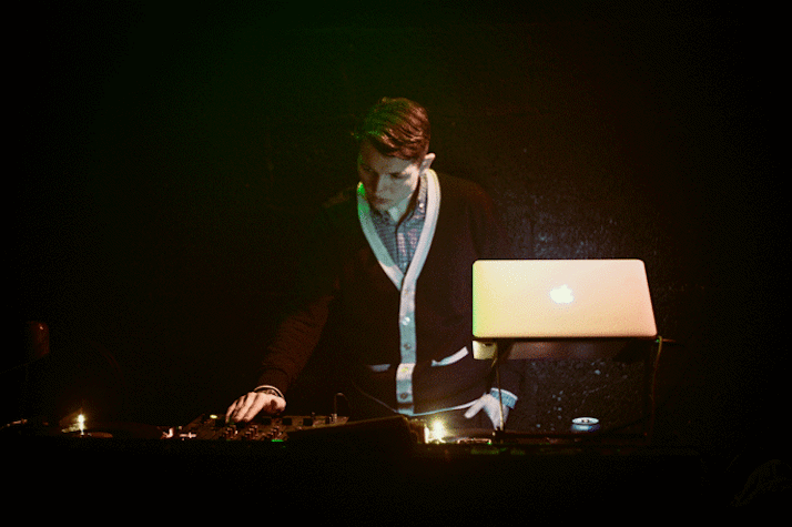 In a dark setting, a man is looking at and working on a DJ console. There is a laptop in front of him.