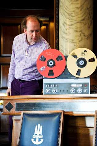 man stood behind a reel to reel tape recorder. infront there is a leather upholstered wooden chair that reads clydeport with a graphic of a trident.