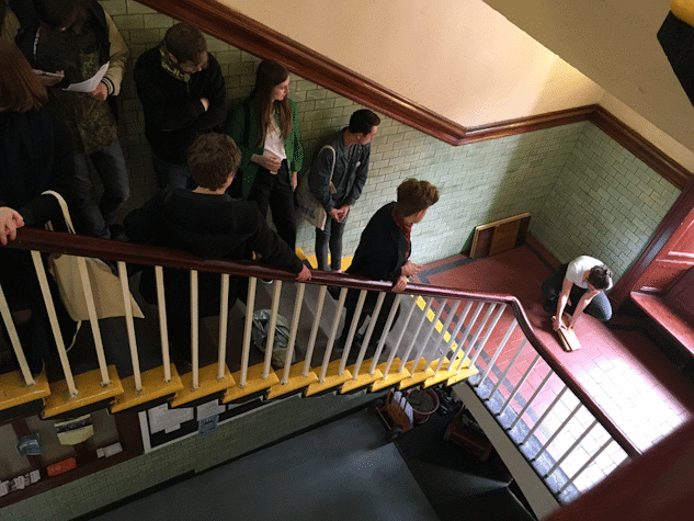 People standing in a stairway looking down at the performance Lost Illusions/Illusions perdues by Sarah Pierce, who has tiles in her hands and is crouched down on the floor.