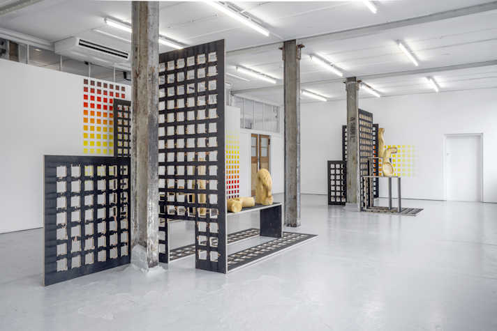 In a large room there are a few wooden sculptures and grid lying vertically and horizontally. The walls have a similar grid painted on them. There are three poles running through the middle of the room and artwork.