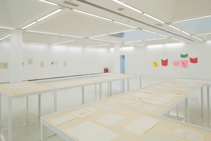 In a large white room, on the wall on the right, six colourful pieces of cloth are hung on the wall. On the left side wall, there are seven paper artworks positioned on the wall. There are two half rectangle shaped large archival tables placed facing away from each other. The tables have various papers and documents placed on them with a generous distance between them.