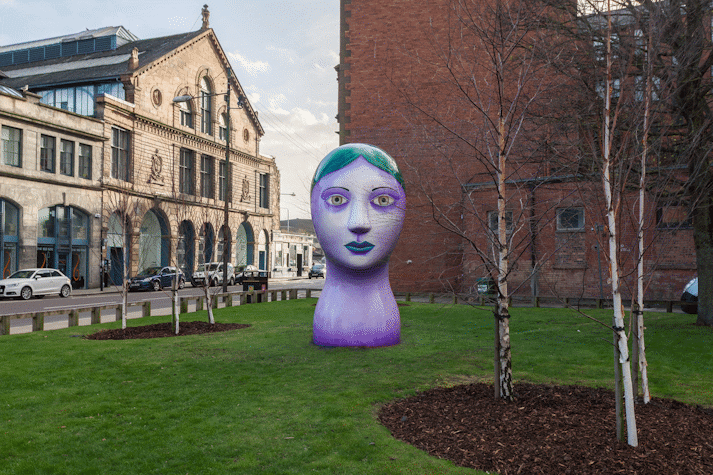Front view of a big, tree-sized bust of a woman with purple skin, green eyes, thin eyebrows and green hair and lips. It is positioned in a park.