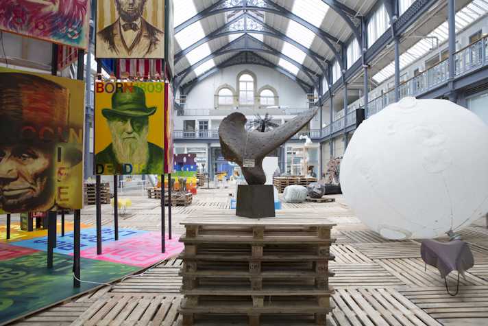 In a large hall, with a glass ceiling, there are multiple sculptures and artworks. On the left, there are iron stands holding posters stacked on top of each other and covering all four sides of the stand. In the centre there is a sculpture and on the right a large blimp like balloon attached to a bicycle seat.