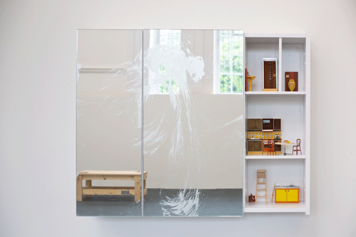 a mirrored cabinet mounted on a wall. One of the doors has been removed and reveals domestic scenes with dolls-house furniture inside.
