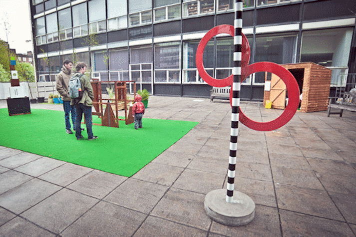 outdoor space with a black and white striped pole with two red circles hanging from it. two adults and a toddler in the background