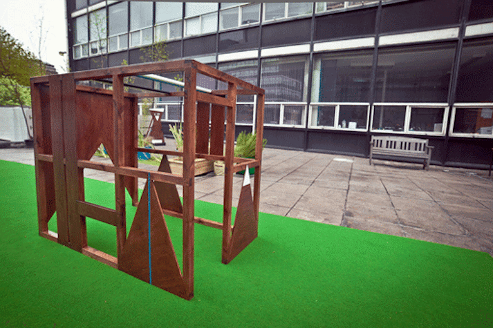 wooden geometric structure on outside on a green rug with a building in the background