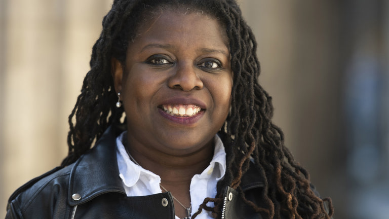 A woman with long black braids smiles, she is wearing a white shirt with a leather jacket over.