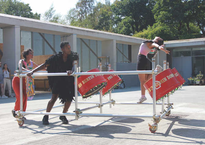Some figures performing with drums wearing bright costumes inside a scaffolding-like structure