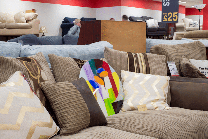 A colorful round painting is lodged in between a brown sofa with brown toned cushions in a British Heart Foundation charity shop.
