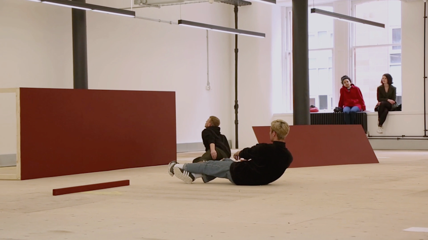 With red panels surrounding them, there are two people in black shirts and blue jeans in movement on the ground. There are two people sitting down in the windowsill, observing.