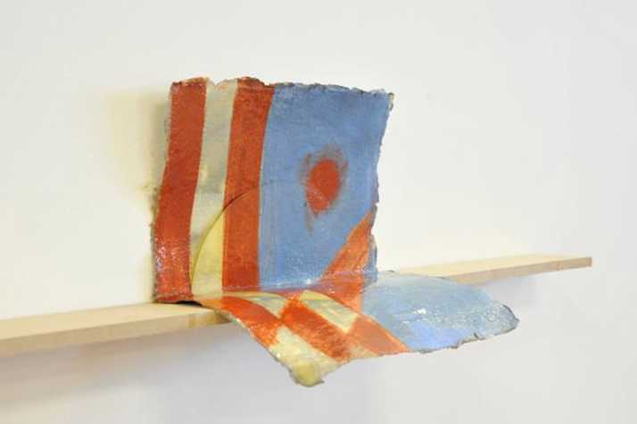 folded painted paper on a small shelf. it has red and grey stripes on one side, and blue with a red dot on the right.