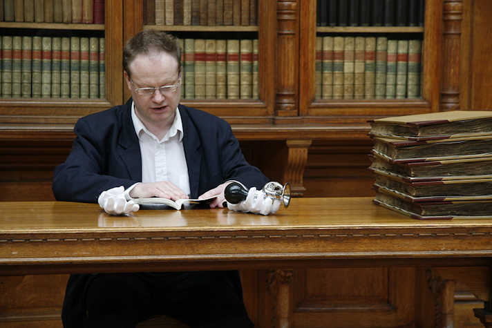 person sat at a wooden desk reading. there is a wall of bookshelves behind. on the desk are two white gloves, one holding a horn