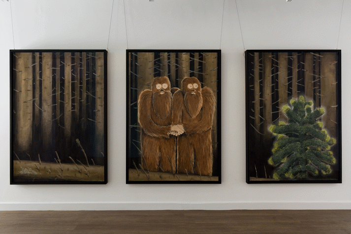 three large scale pastel drawings in black frames and portrait orientation hang on a white wall. they have a shared backdrop of a dark leafless forest. the left drawing shows tree trunks, the centre drawing shows two yeti figures embracing one another, the right drawing shows a monkeypuzzle tree.